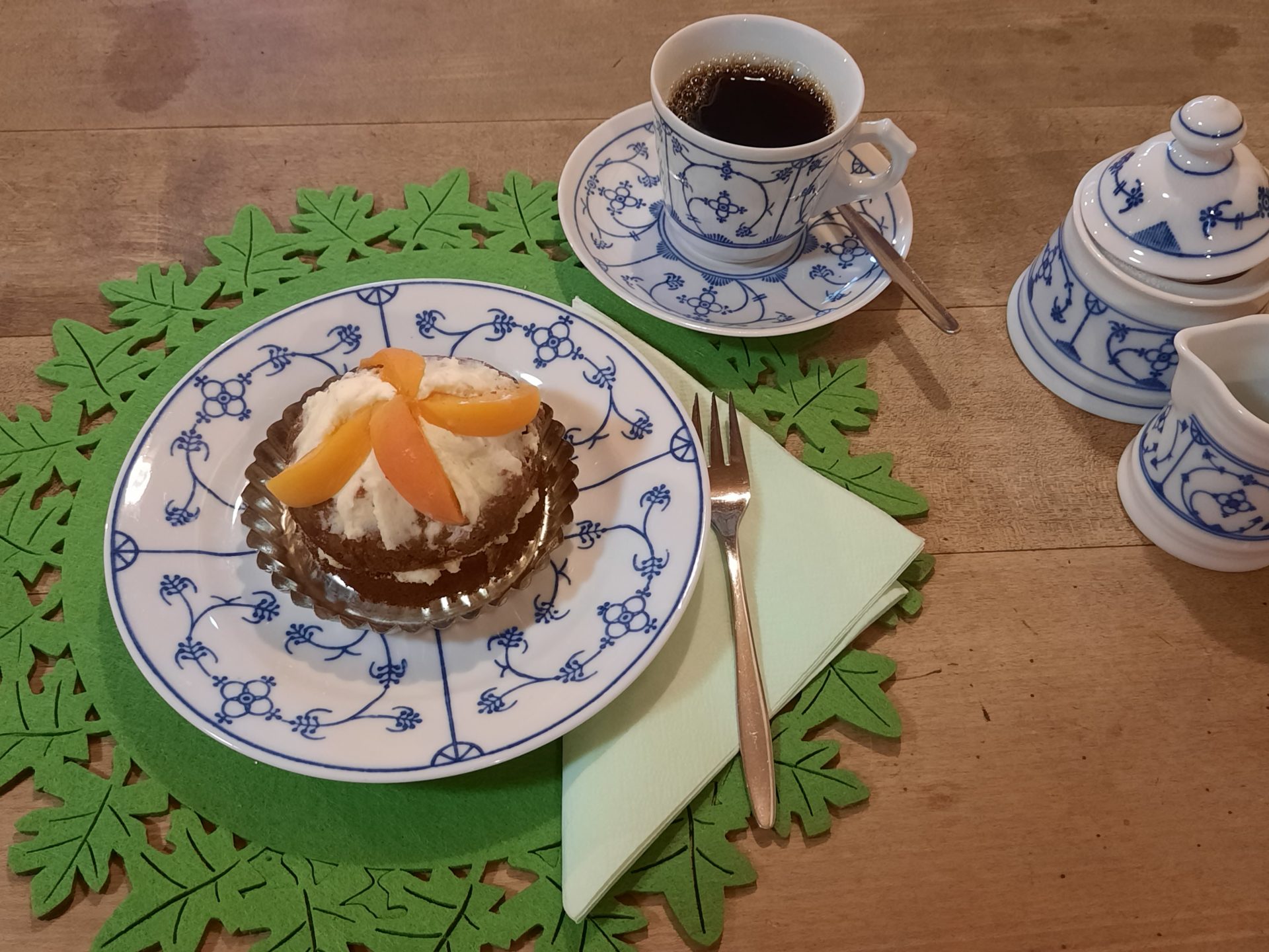 Ein Aprikosentörtchen und eine Tasse Kaffee auf Zwiebelmuster-Geschirr, dazu Sahnekännchen und Zuckerdose auf grüner Filzunterlage.