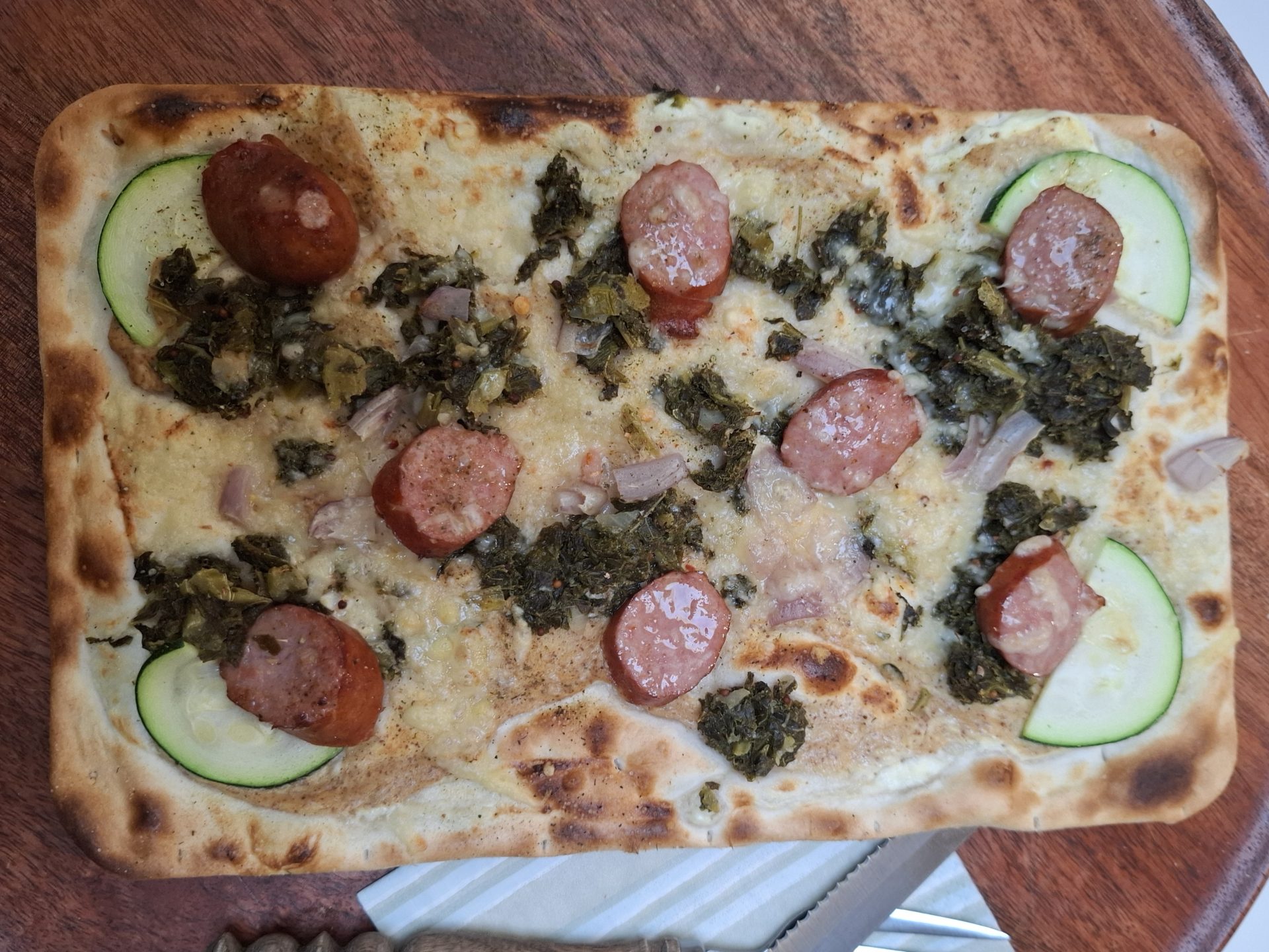 Ein Flammkuchen mit Mettwurstscheiben, Grünkohl und Zucchini auf einem runden Holzteller, daneben Messer und Gabel.
