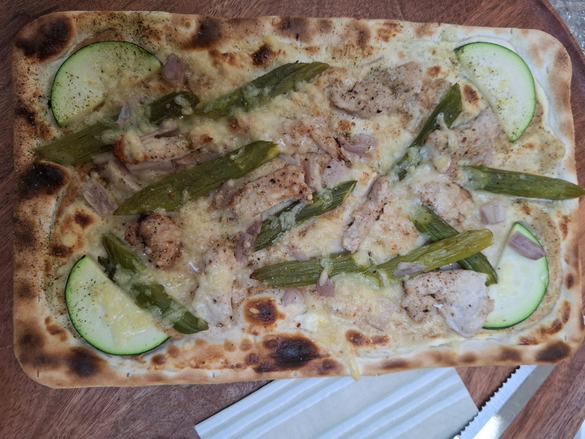 Ein Flammkuchen mit grünem Spargel, gebratenem Fleisch, Schinken und Zucchini auf einem Holzteller.