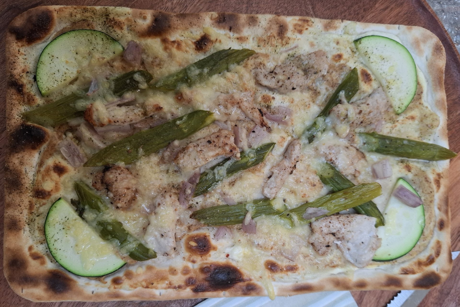 Ein Flammkuchen mit grünem Spargel, gebratenem Fleisch, Schinken und Zucchini auf einem Holzteller.