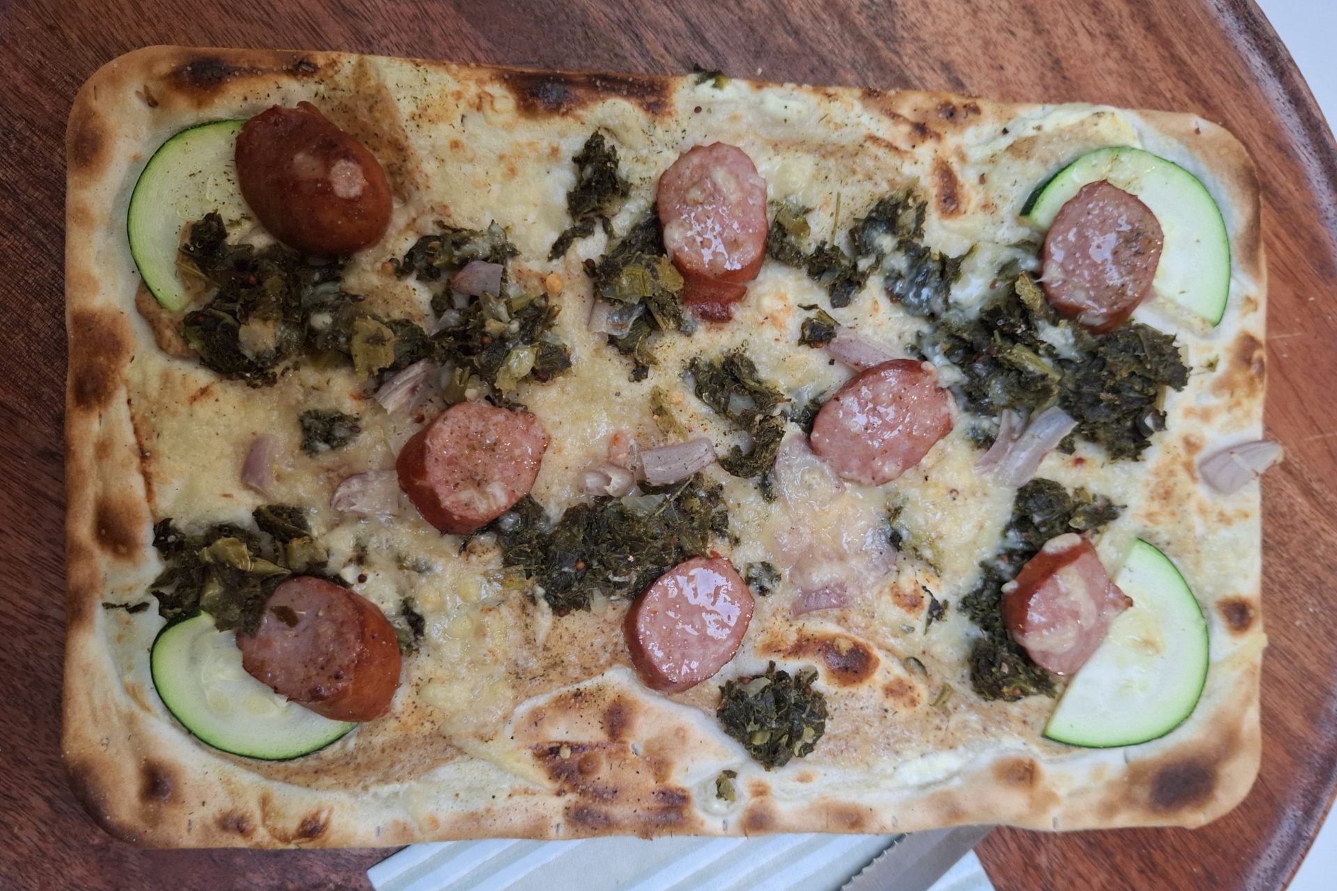 Ein Flammkuchen mit Mettwurstscheiben, Grünkohl und Zucchini auf einem runden Holzteller, daneben Messer und Gabel.