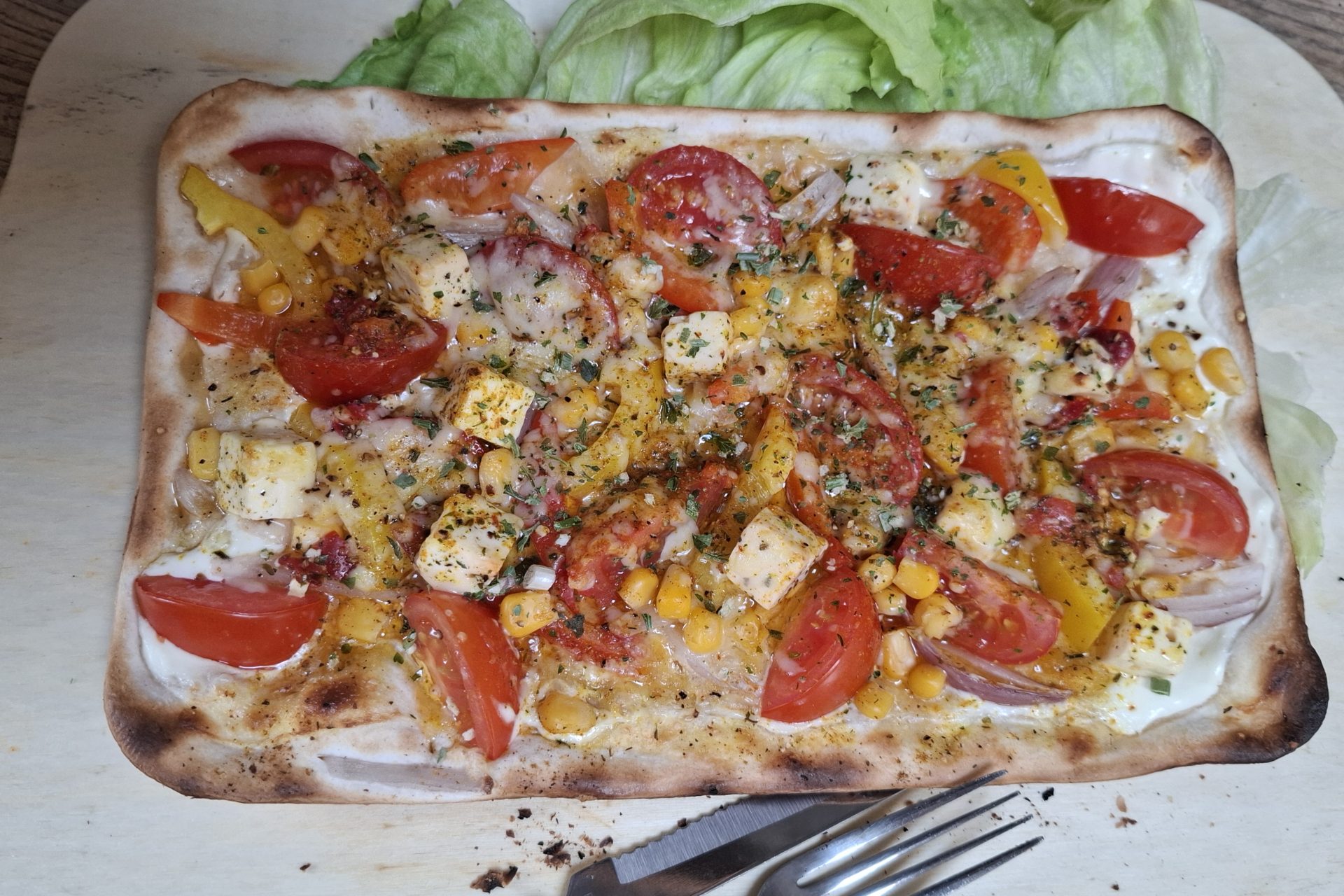 Ein Flammkuchen mit Tomaten, Feta, Mais und Zwiebeln auf einem hölzernen Servierbrett, daneben Blattsalat und Besteck.
