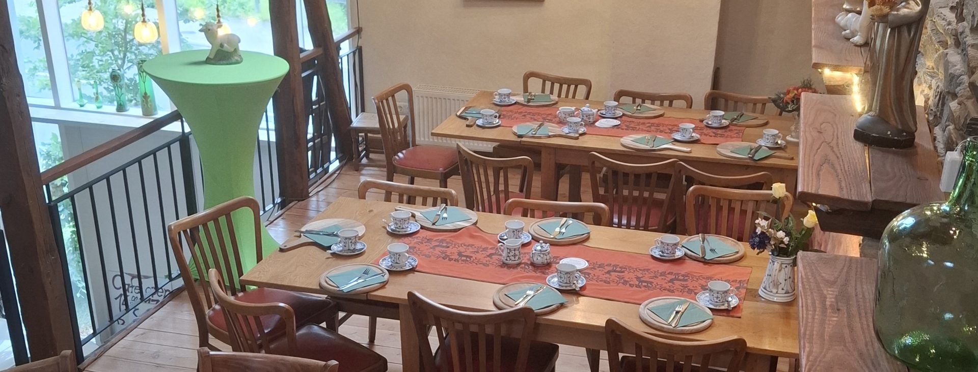 Die Festhalle im alten Heustall, für eine Kaffeetafel gedeckt: lange Tische mit Tischläufern und Gedecken unter dem Kronleuchter, links die Galerie zum Fenster.