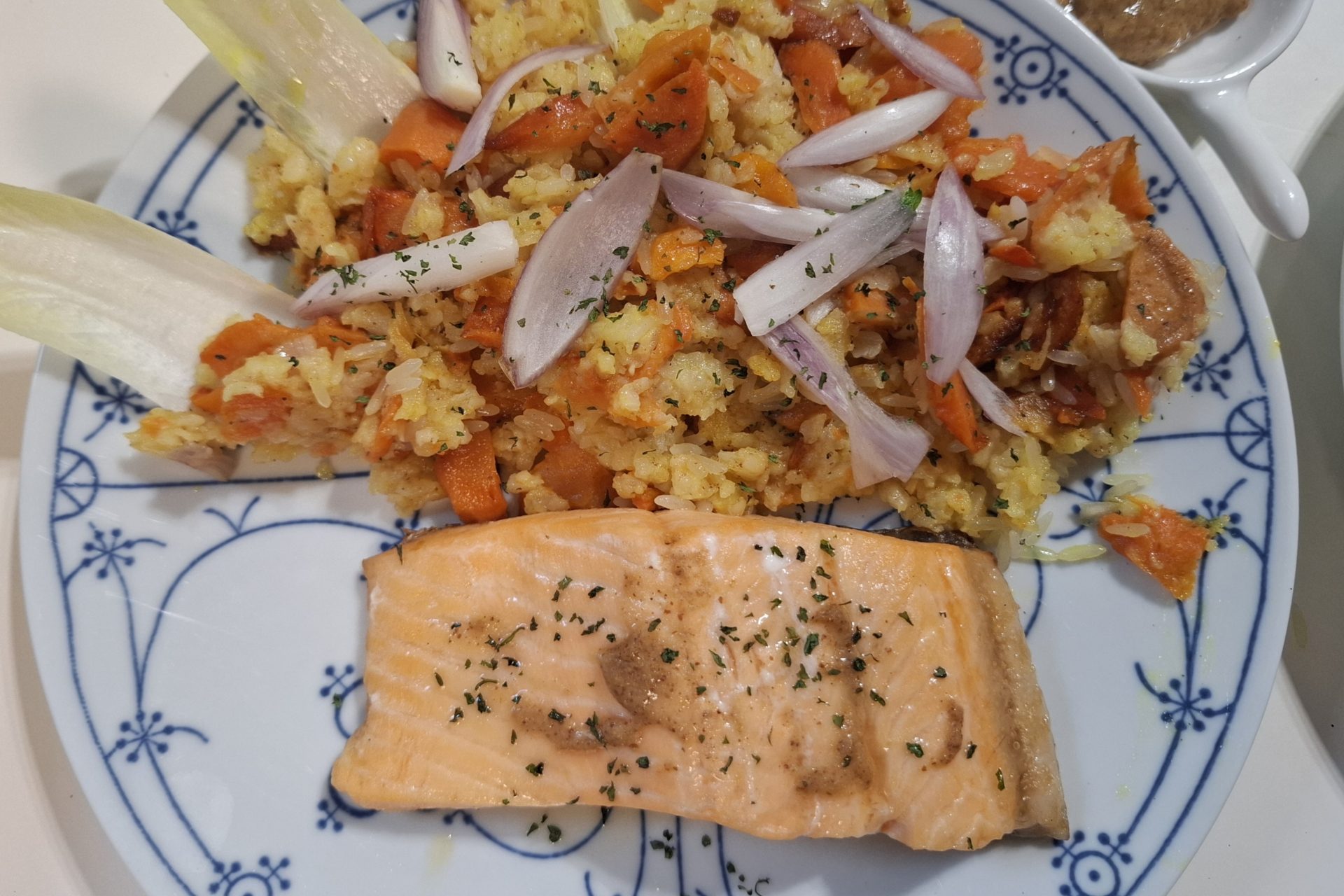 Ein Lachsfilet auf Gemüsereis, mit Chicorée-Blättern und Zwiebelringen angerichtet, auf blau-weißem Zwiebelmuster-Teller.