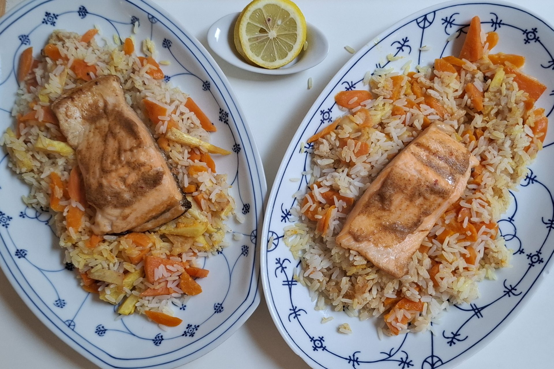 Zwei Portionen gebratenes Lachsfilet auf Karotten-Reis, auf ovalen Zwiebelmuster-Platten, daneben eine halbe Zitrone.