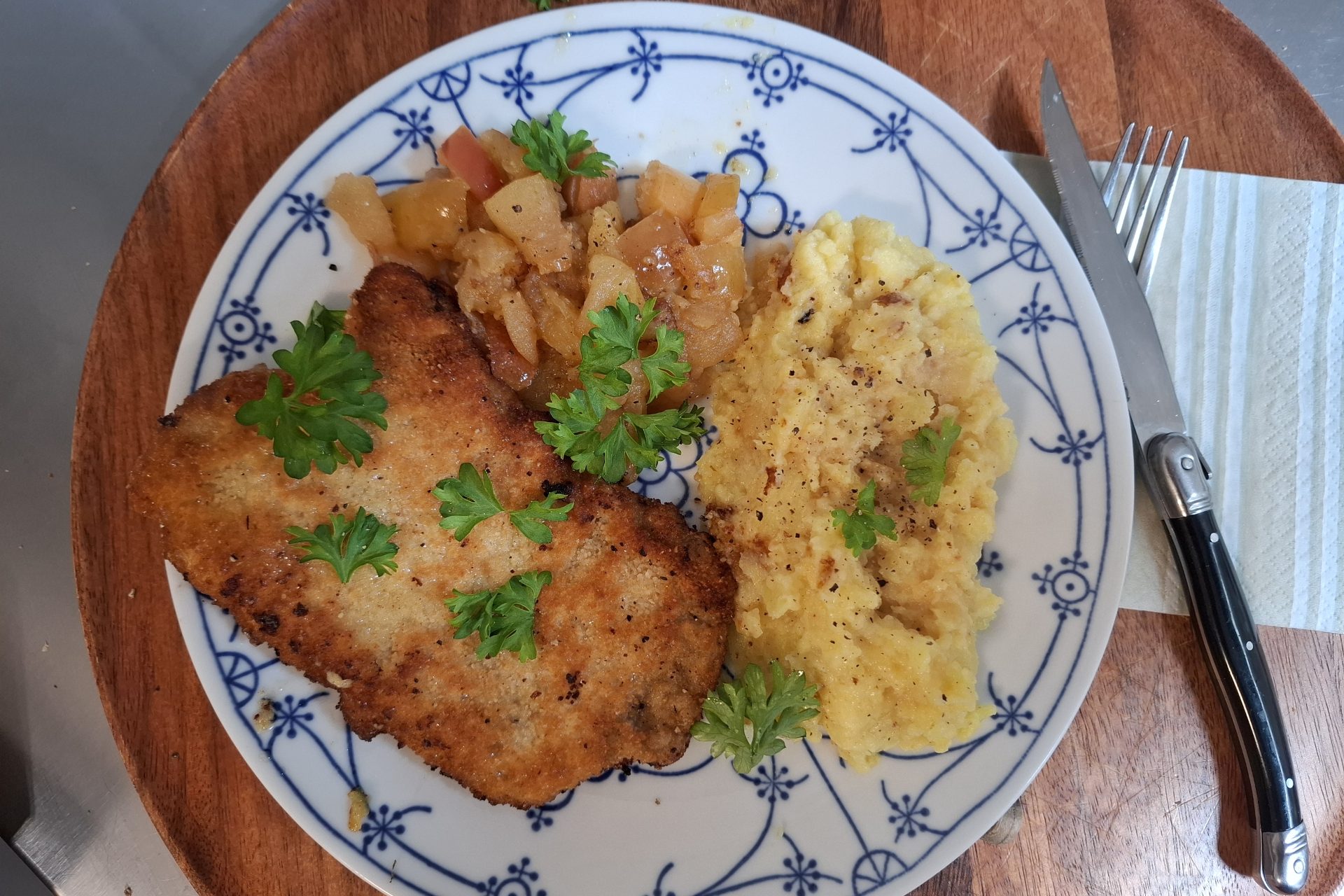 Ein paniertes Schnitzel mit Kartoffelstampf und Apfelkompott, mit Petersilie angerichtet.