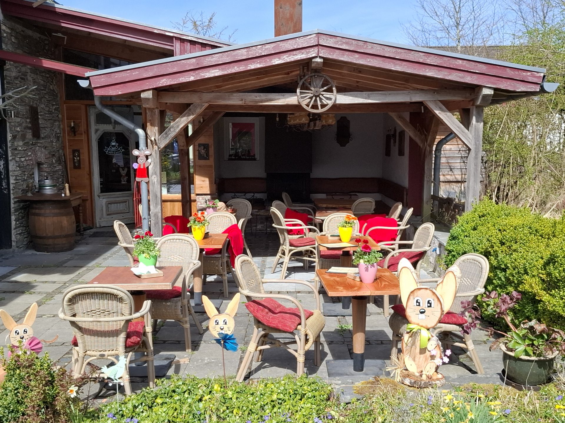 Die Terrasse unter der hölzernen Pergola, Korbstühle mit roten Kissen an gedeckten Tischen, davor Osterhasen aus Holz und blühende Frühlingsbeete.