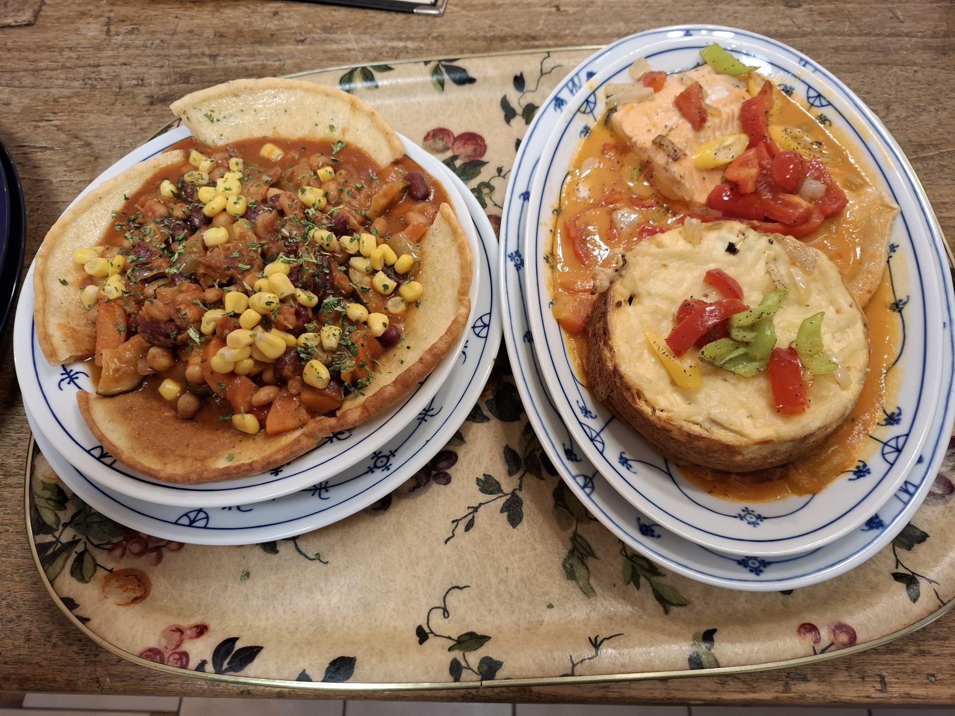 Zwei Gerichte auf einem Tablett: ein Fladen mit Chili con Carne und Mais, daneben ein Lachsfilet mit Paprika und ein überbackenes Brot.