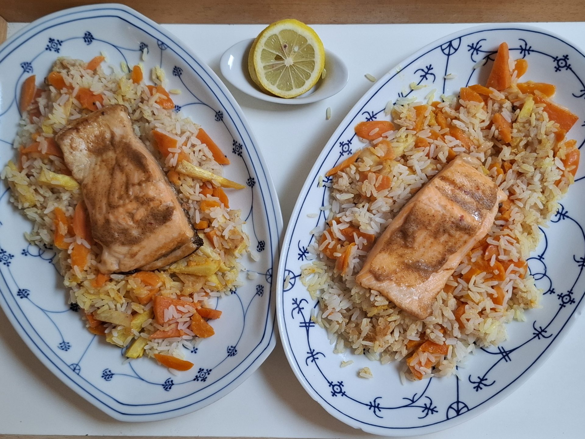 Zwei Portionen gebratenes Lachsfilet auf Karotten-Reis, auf ovalen Zwiebelmuster-Platten, daneben eine halbe Zitrone.