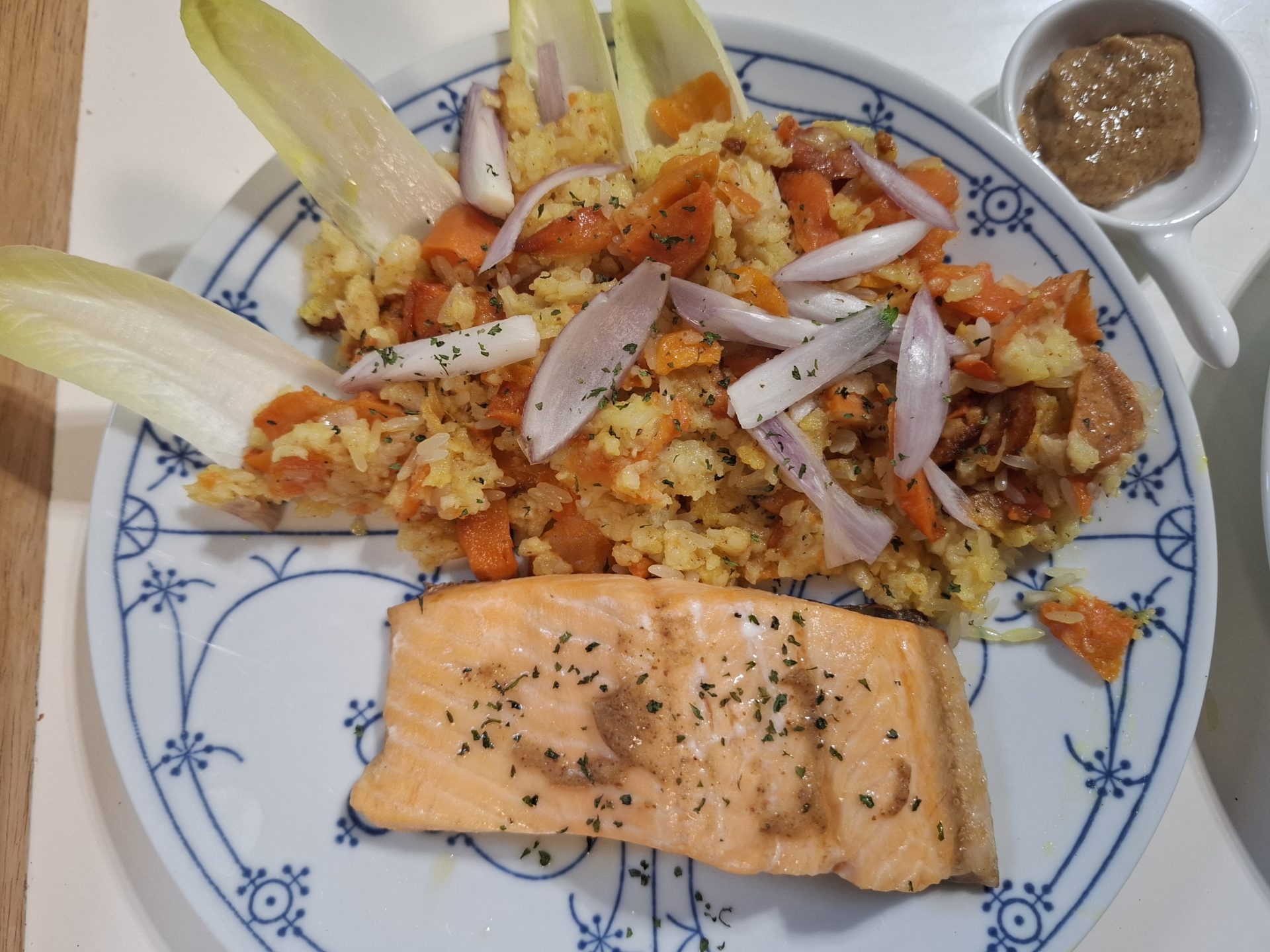 Ein Lachsfilet auf Gemüsereis, mit Chicorée-Blättern und Zwiebelringen angerichtet, auf blau-weißem Zwiebelmuster-Teller.